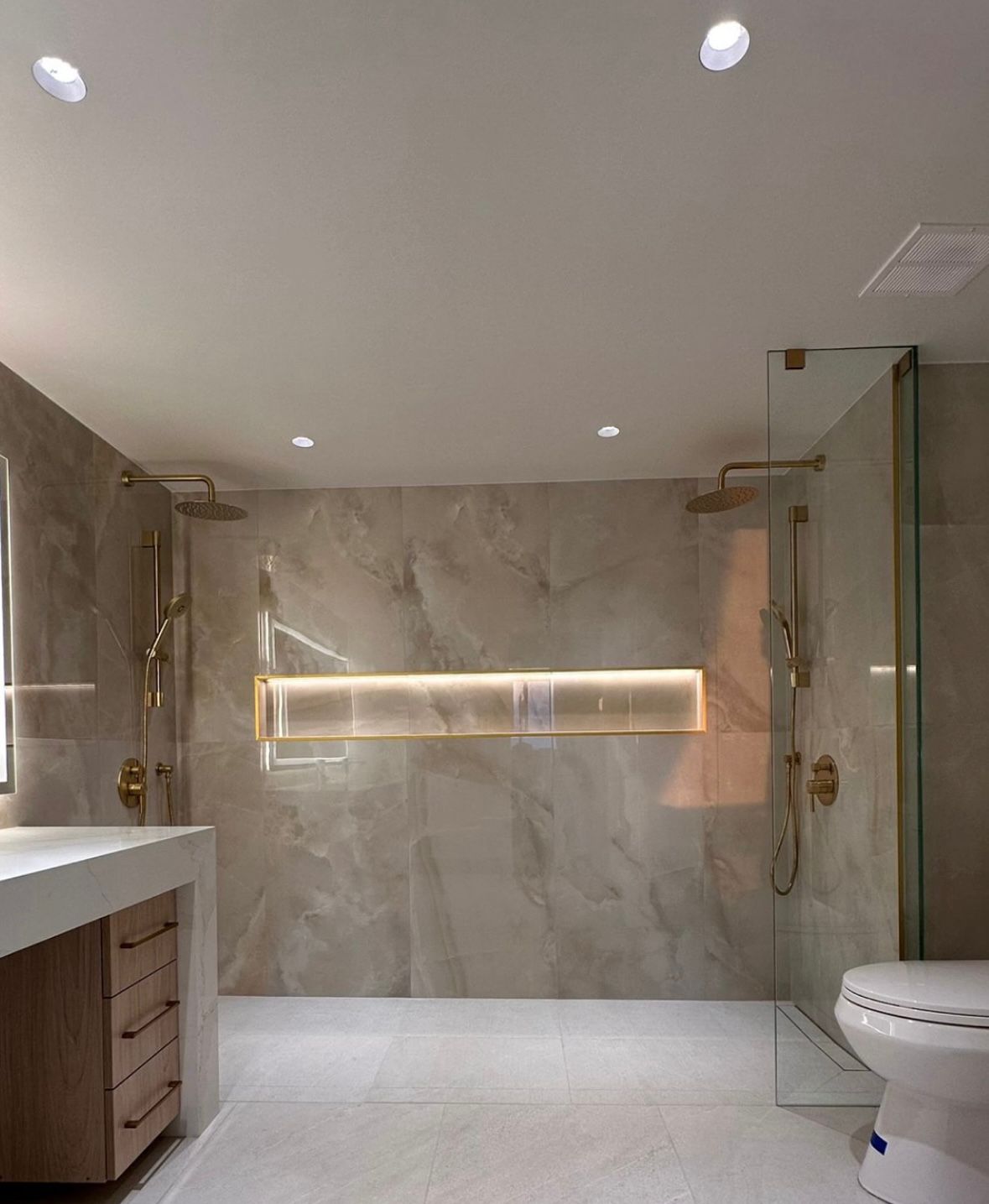 Large curbless double walk-in shower with two gold rainfall heads, a glass screen and a backlit niche